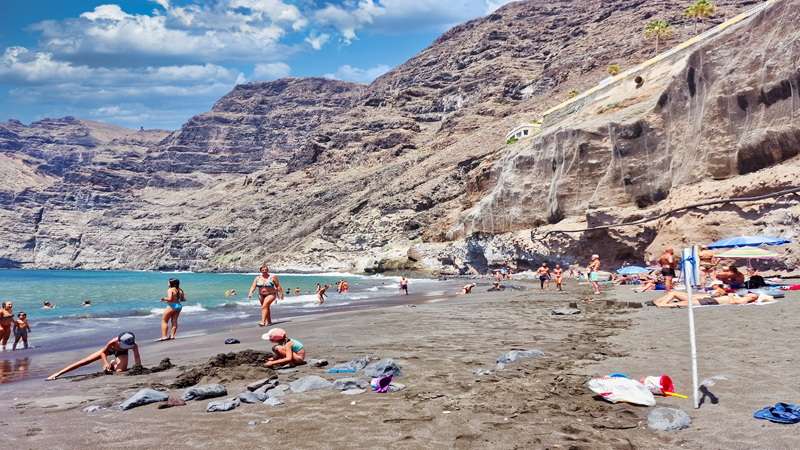 plaża z ciemniejszym piaskiem pełna turystów przy skałach Los Gigantes na Teneryfie